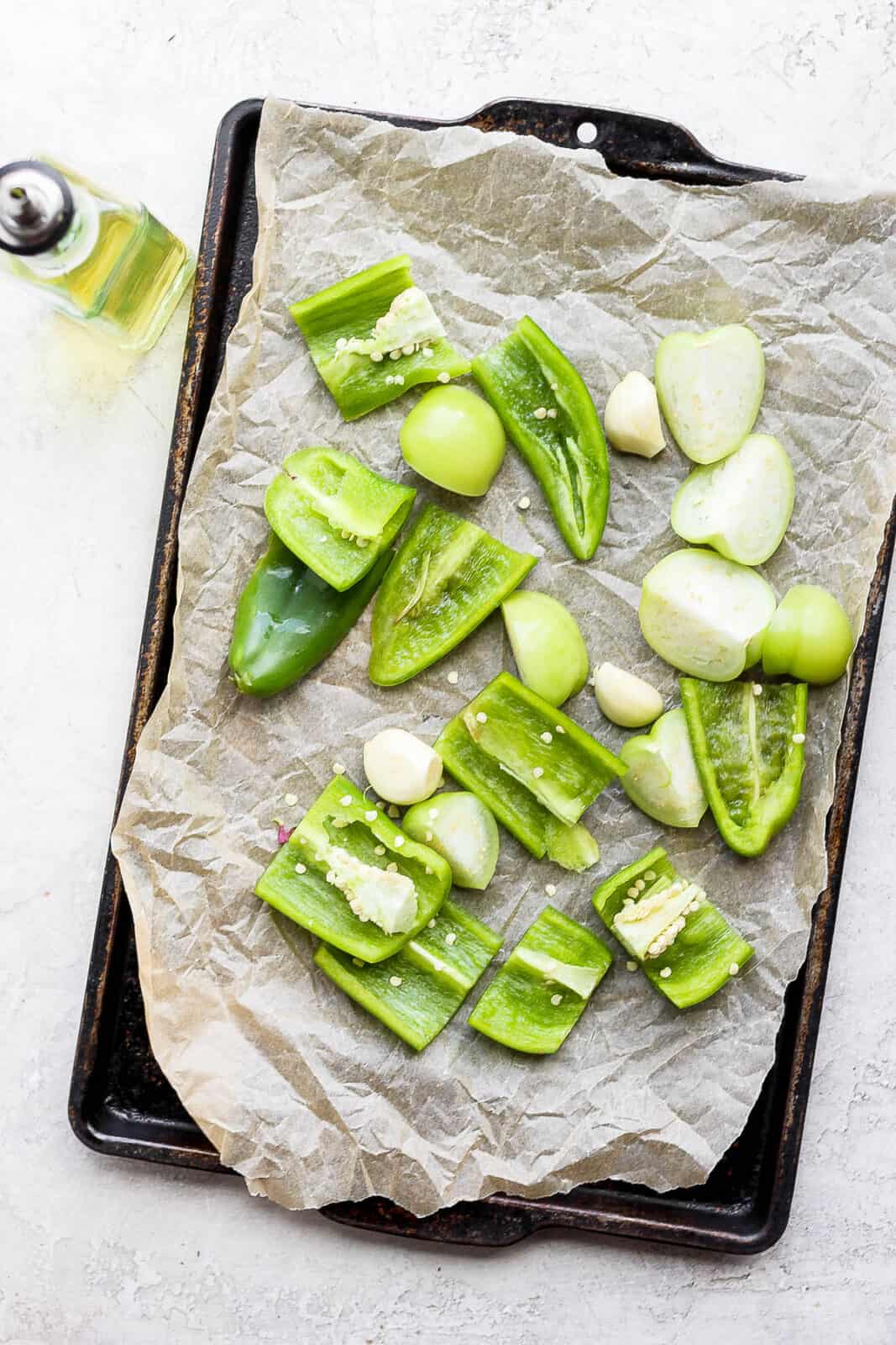 Tomatillos, anaheim pepper, jalapeno, garlic, and onion on a parchment-lined pan with olive oil next to it.