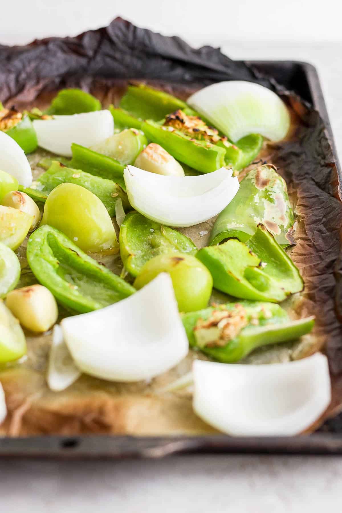 Roasted tomatillos, anaheim pepper, jalapeno, garlic, and onion on a parchment-lined pan.