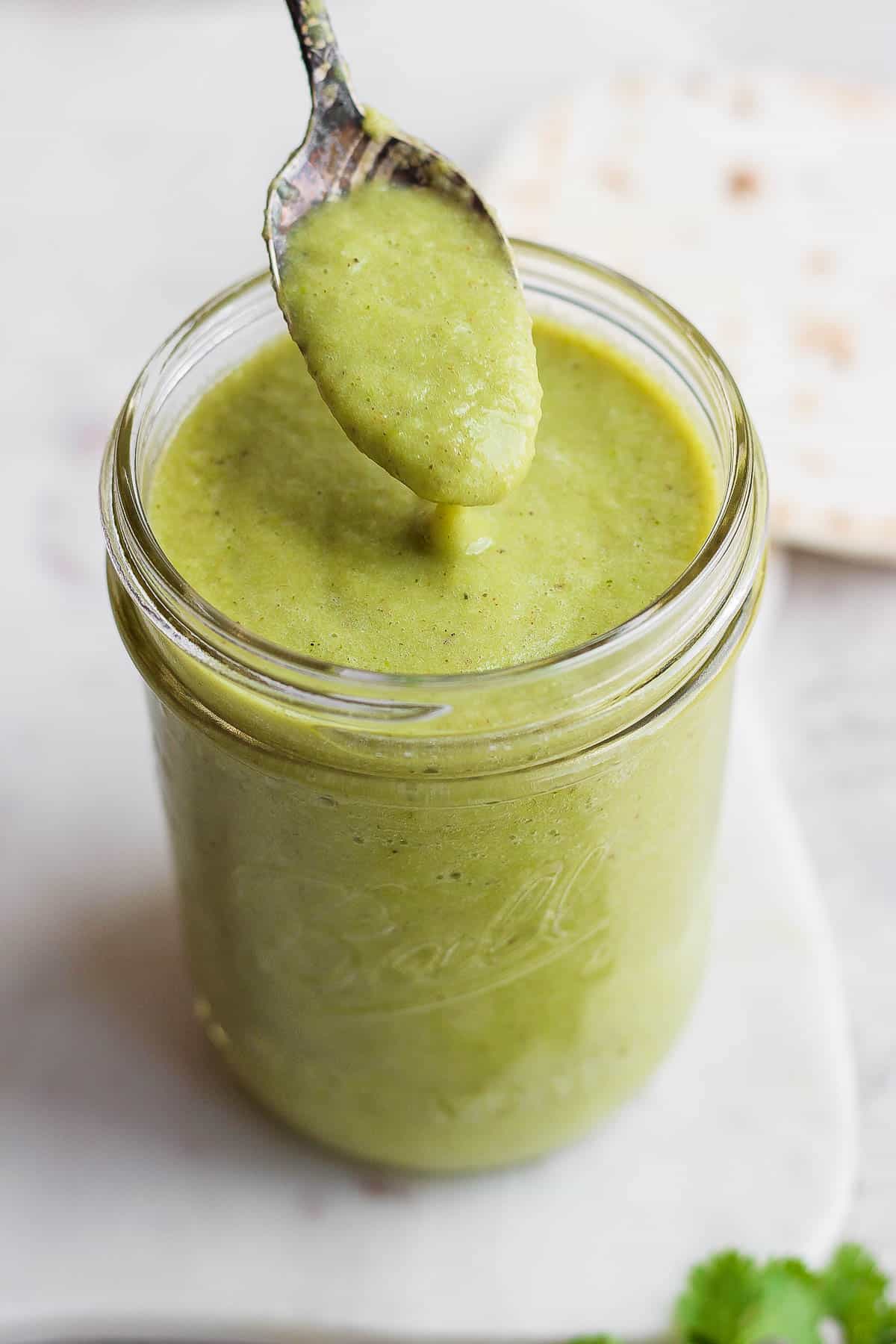 A spoon scooping green enchilada sauce in a mason jar.