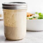 Jar of homemade caesar dressing in a mason jar with lid on and salad in a bowl behind it.