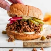 A hand placing the top bun on a sloppy joe topped with red onions and pickles.