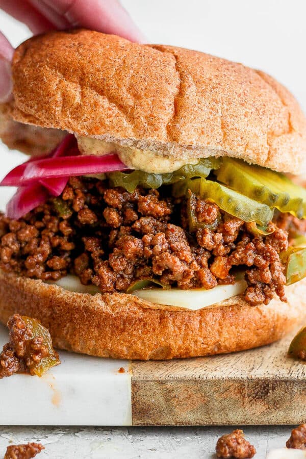 A hand placing the top bun on a sloppy joe topped with red onions and pickles.