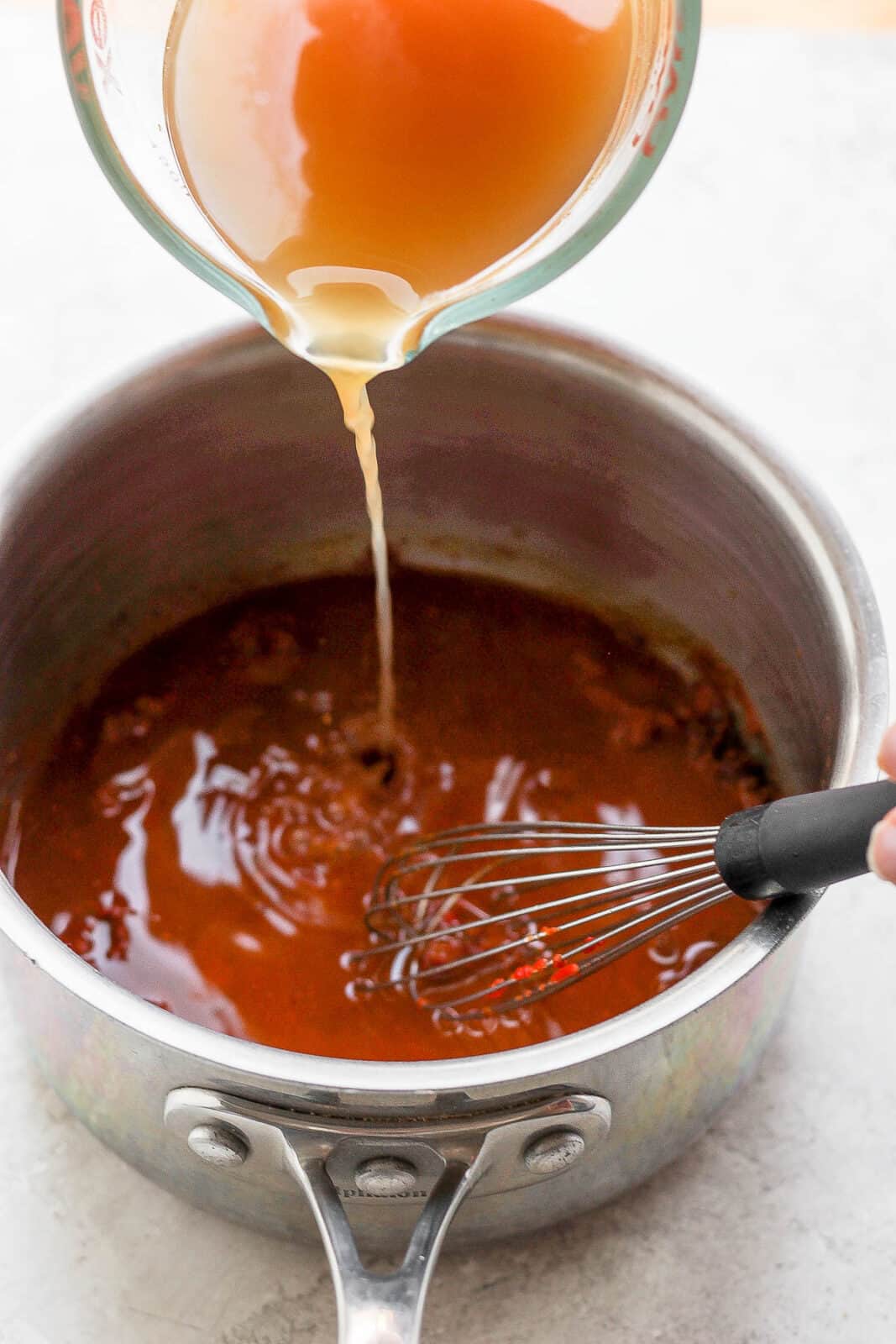 Chicken broth being poured into enchilada sauce in a saucepan and a whisk.