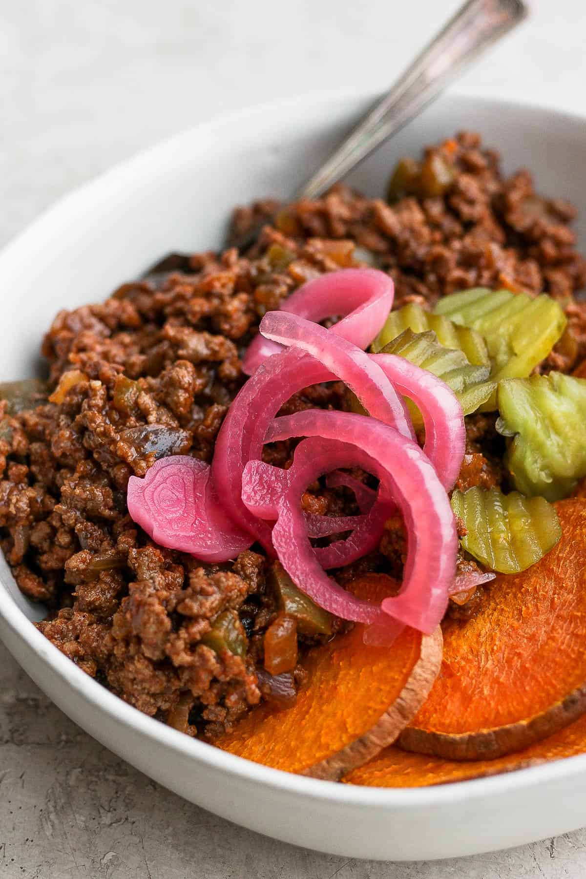A sloppy joe bowl with pickled red onions and burger pickles on top.