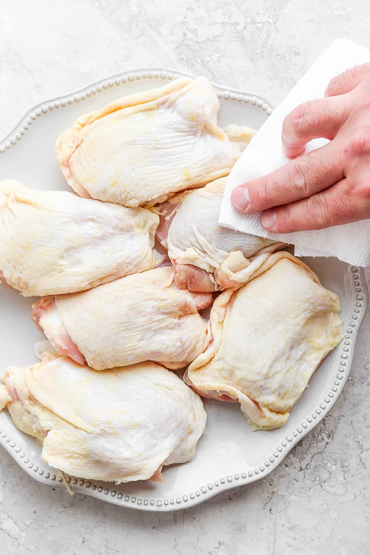 A plate of chicken thighs with someone patting them dry with a clean paper towel.