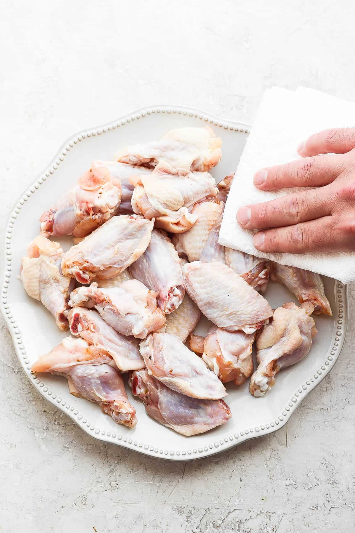 Raw chicken wings on a plate being patted dry with a paper towel. 