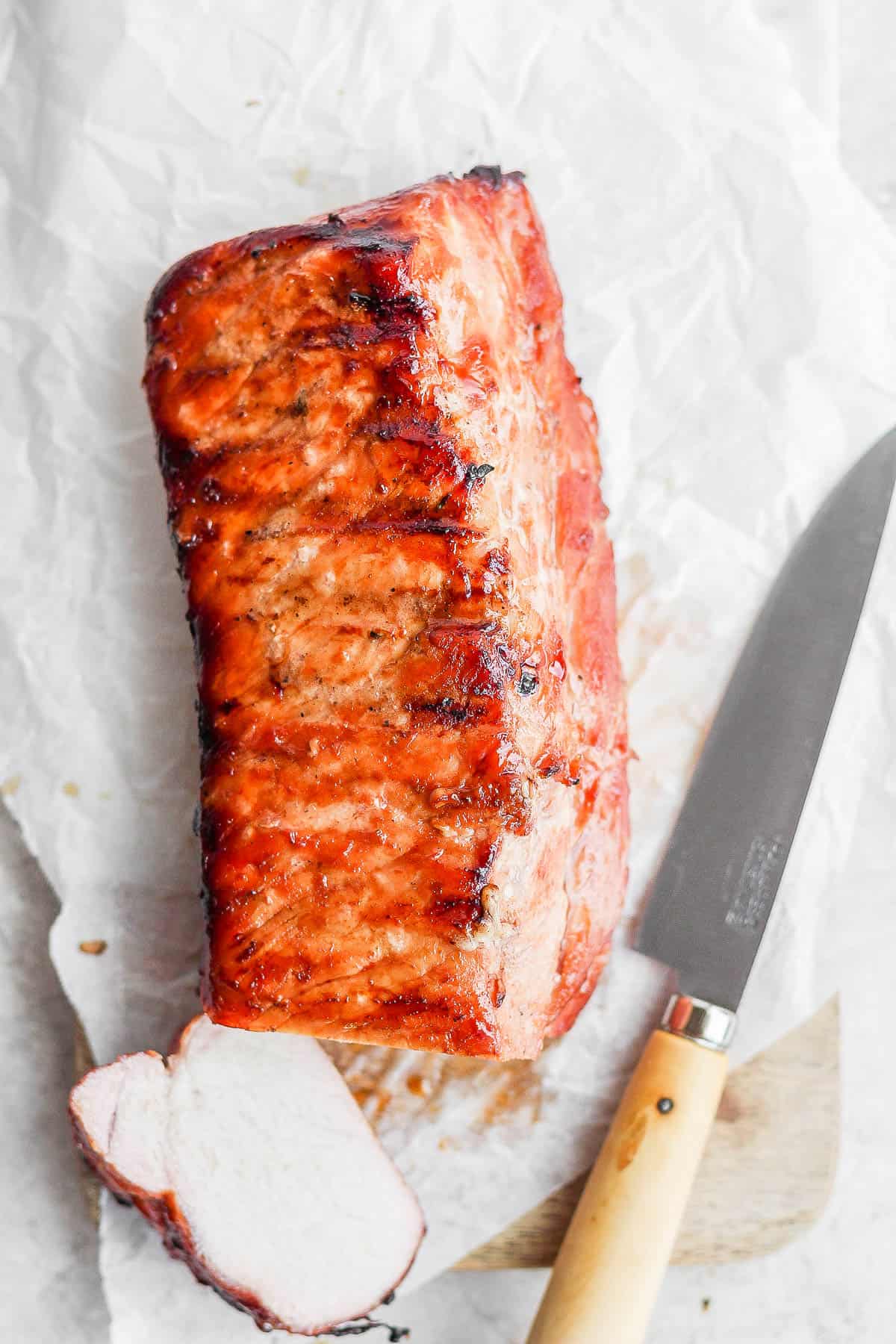 A pork loin on a wooden board with a knife next to it and the end cut off. 