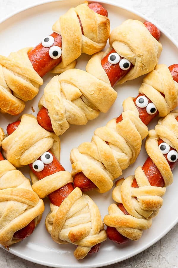 Six mummy hot dogs on a white plate with candy eyes.