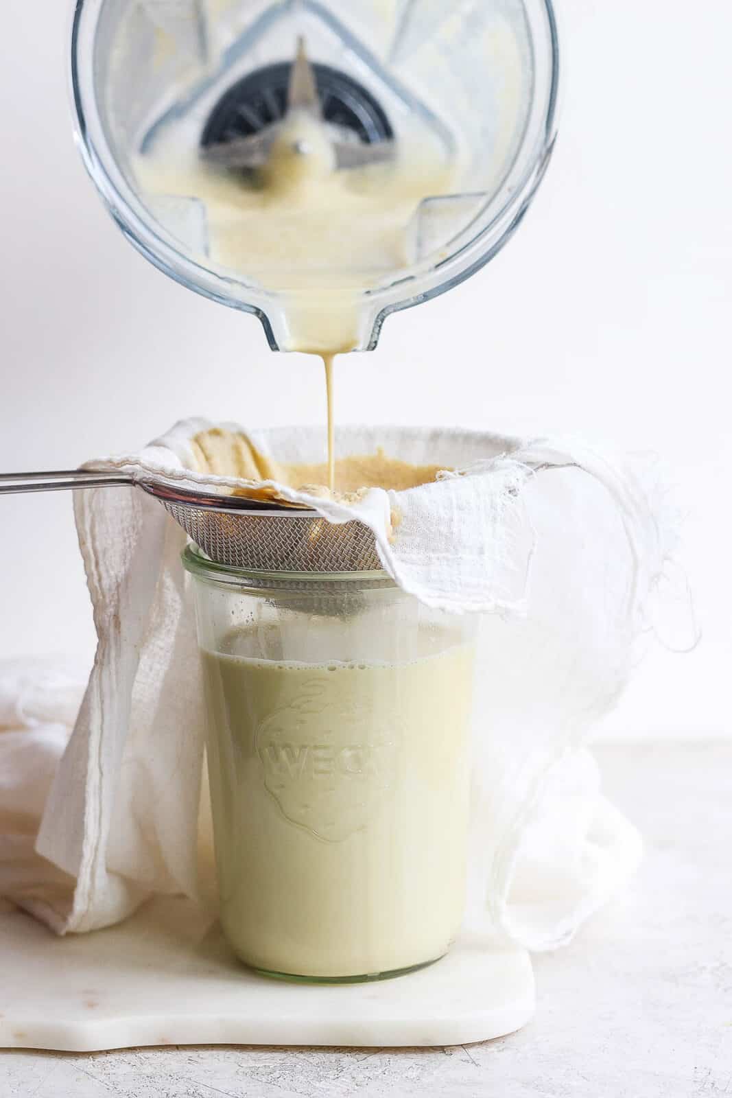 Pouring pistachio milk through a cheesecloth and sieve and into a large jar.