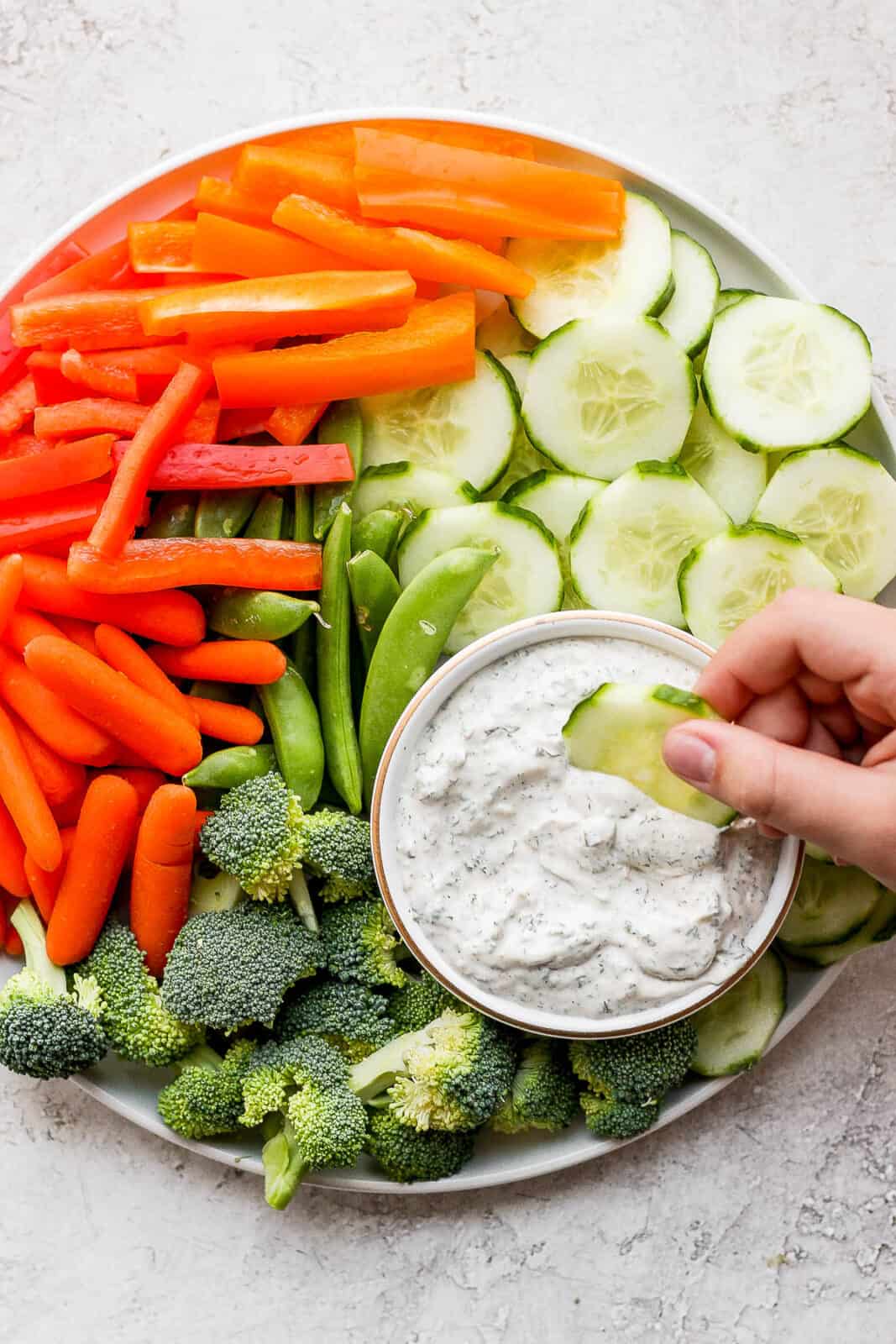 A cucumber being dipped in a small dish of healthy veggie dip on a plate of veggies.