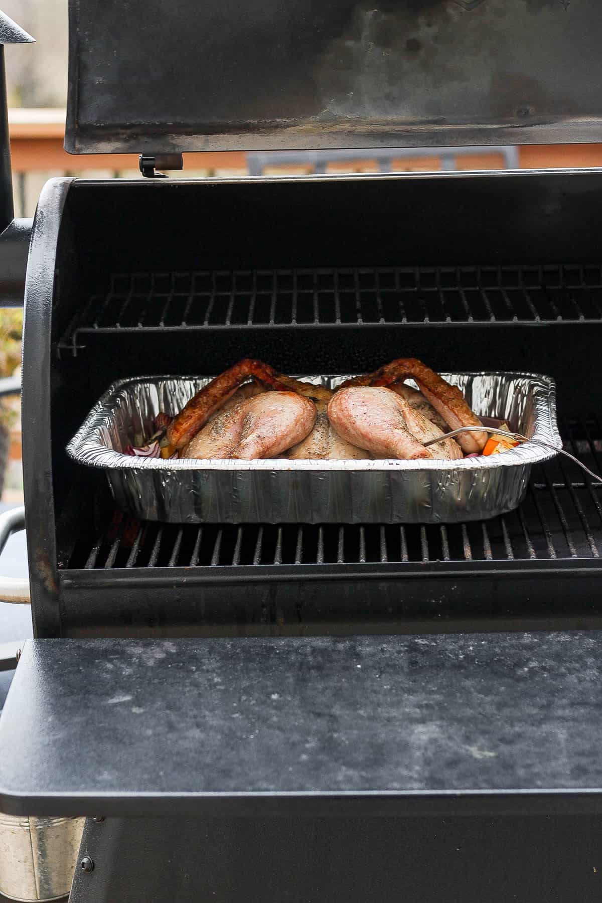 A spatchcocked turkey in an aluminum roasting pan on a smoker.