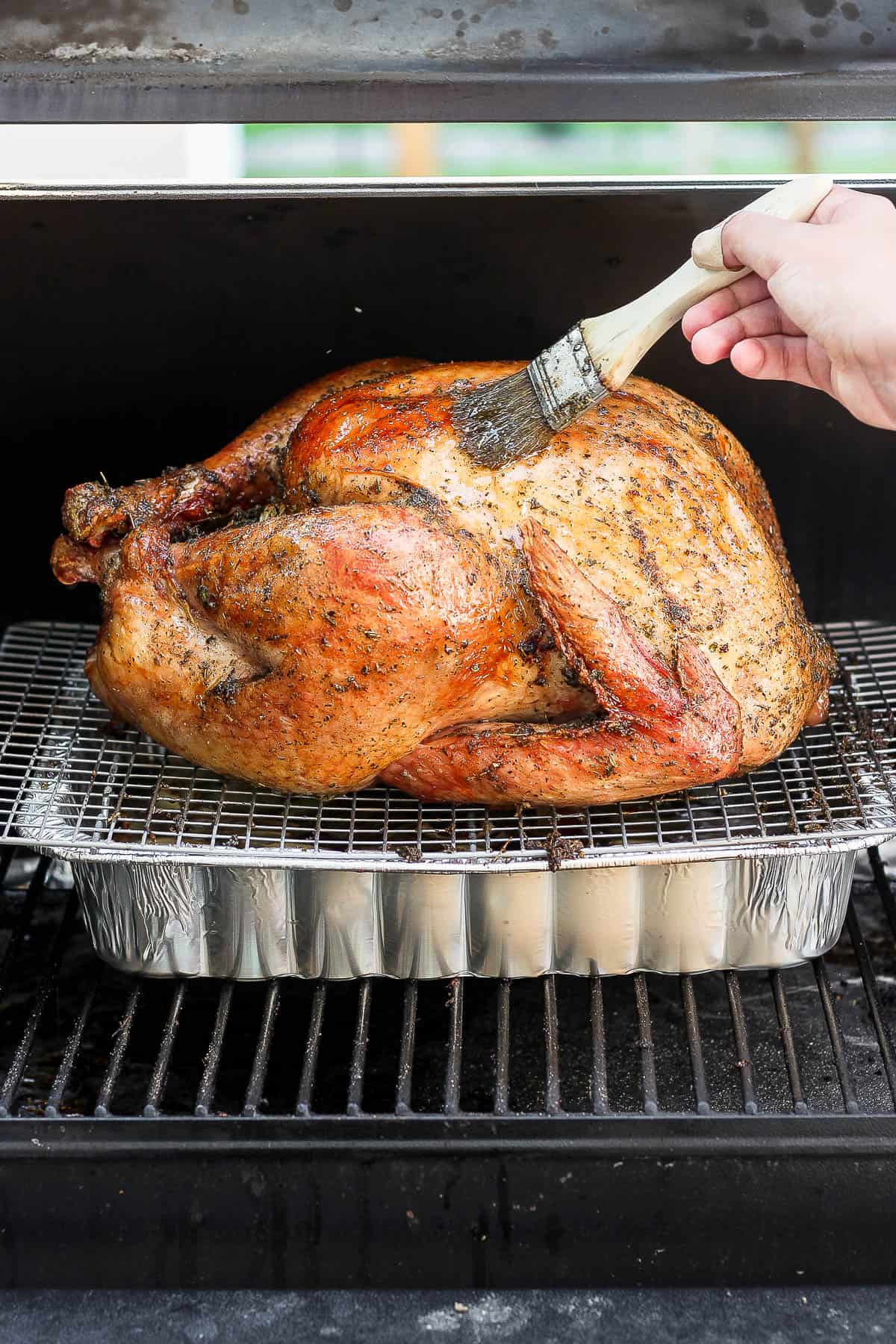 A whole turkey being cooked on a smoker with someone basting it with a brush. 