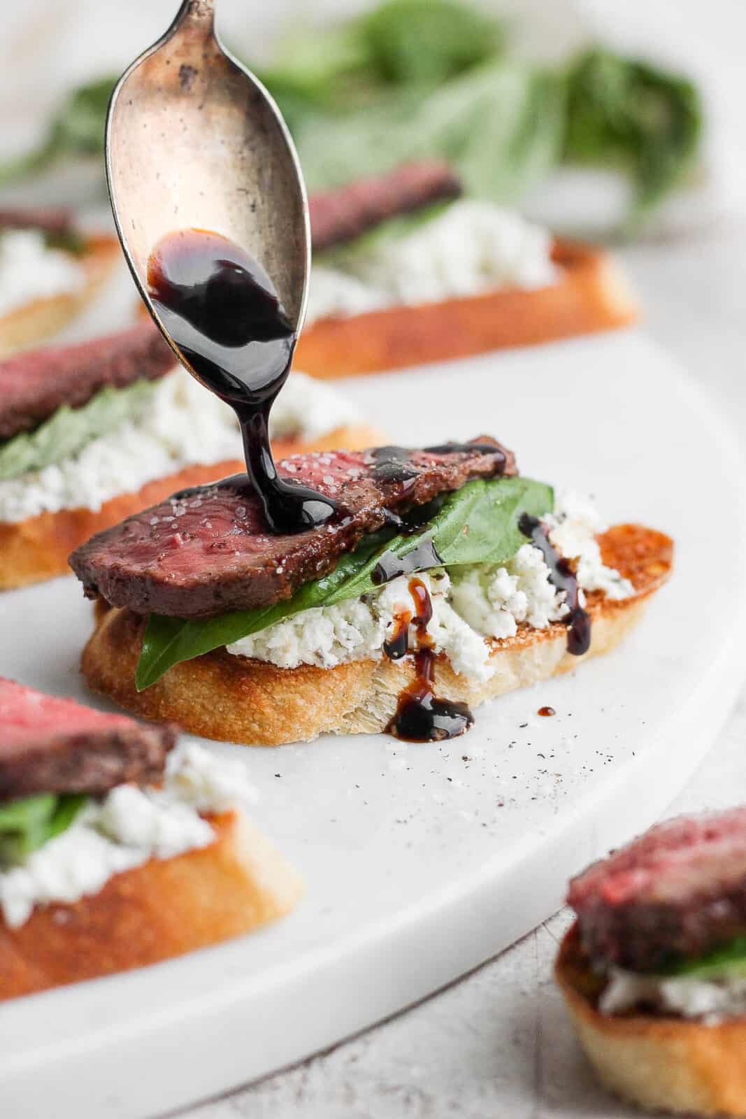A steak crostini with goat cheese, basil and someone drizzling balsamic glaze. 