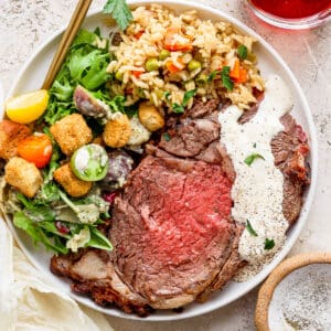 Top down shot of a plate of prime rib with horseradish sauce on top, rice pilaf and a house salad.