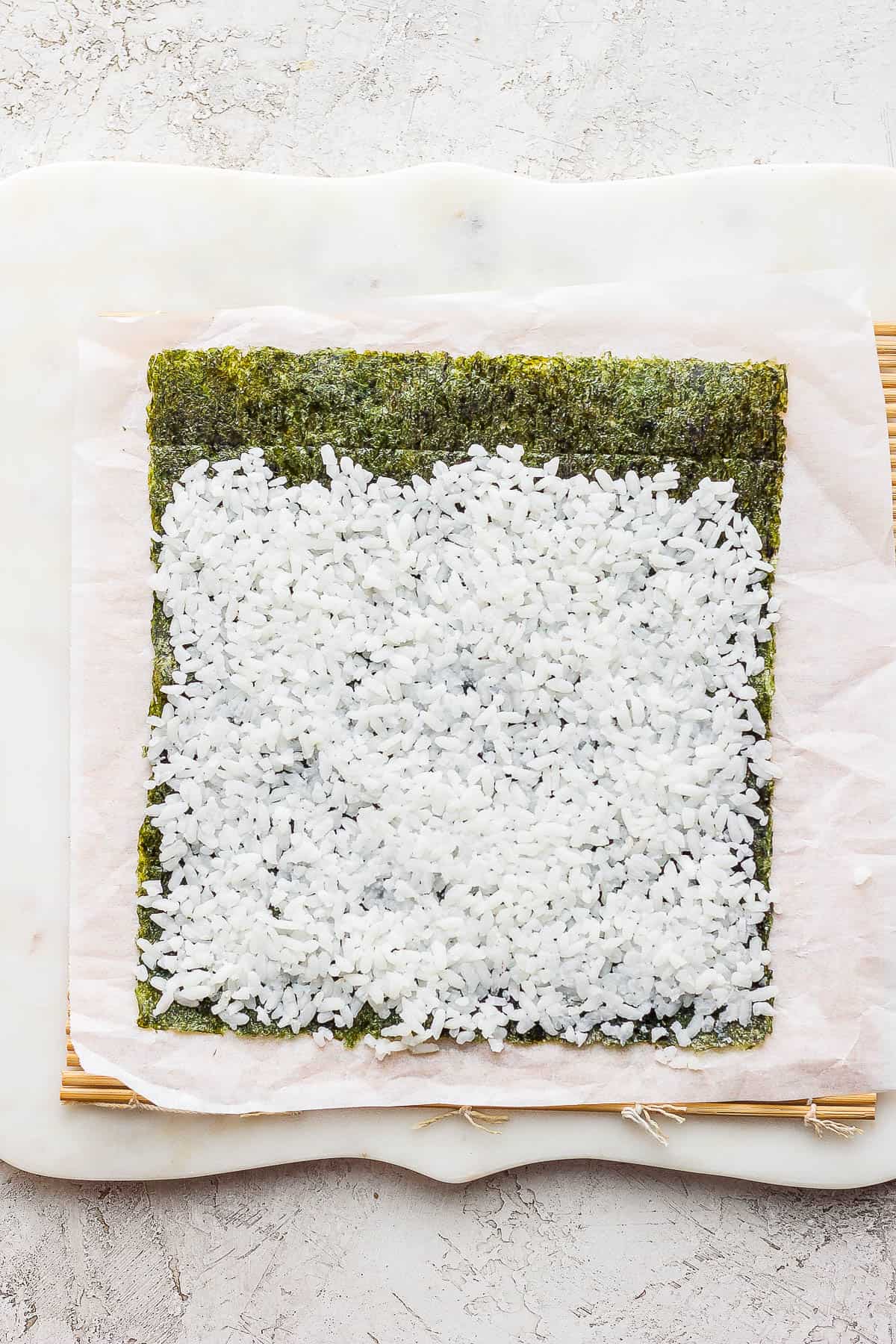 A nori sheet on a parchment lined bamboo sheet with rice spread on top of it.