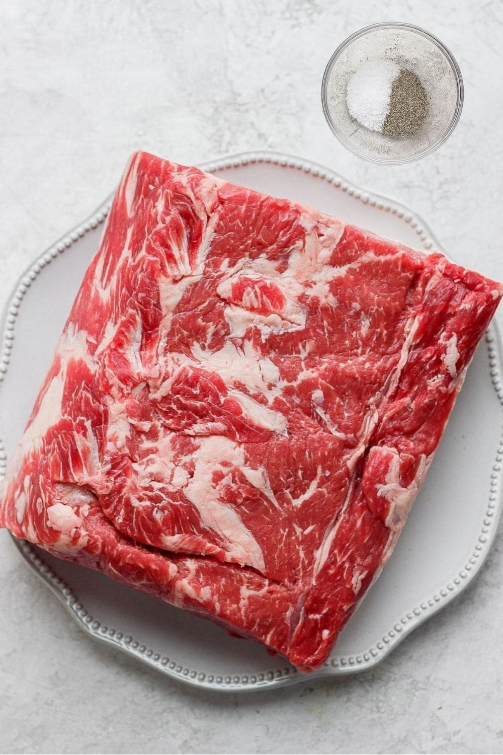 A raw standing rib roast on a plate and small bowl with kosher salt and ground black pepper in it. 