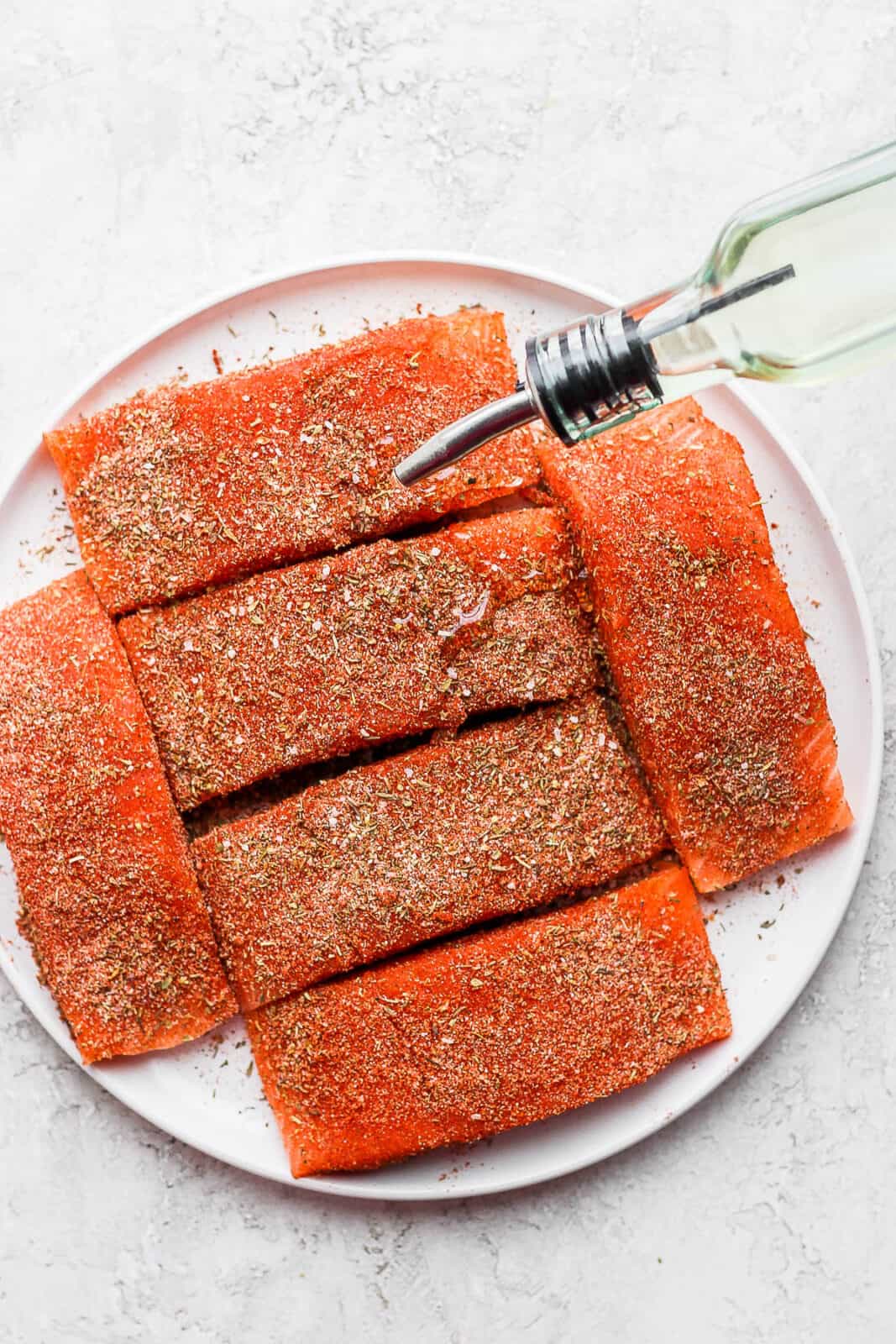 Olive oil being drizzled on seasoned salmon fillets on a plate.