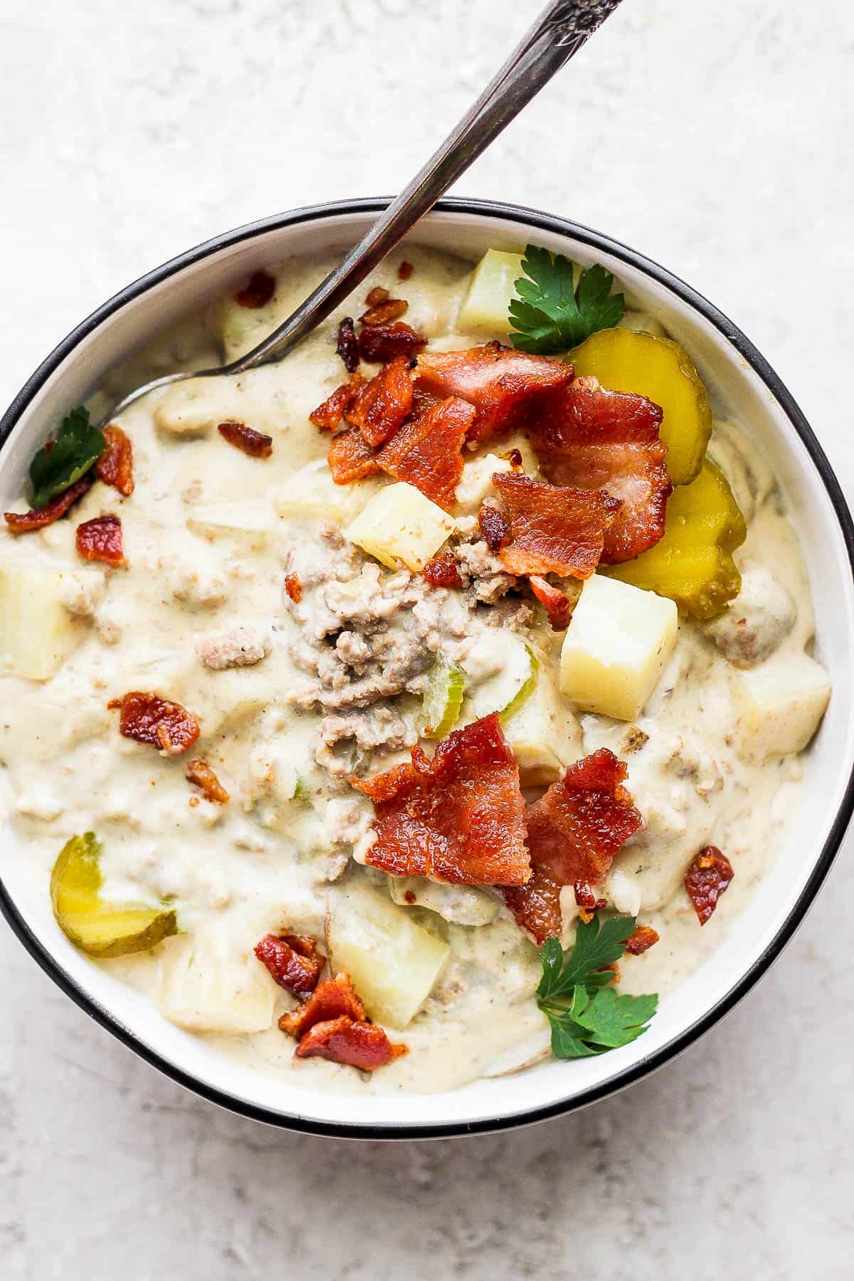 Bowl of cheeseburger soup garnished with pickles and bacon.