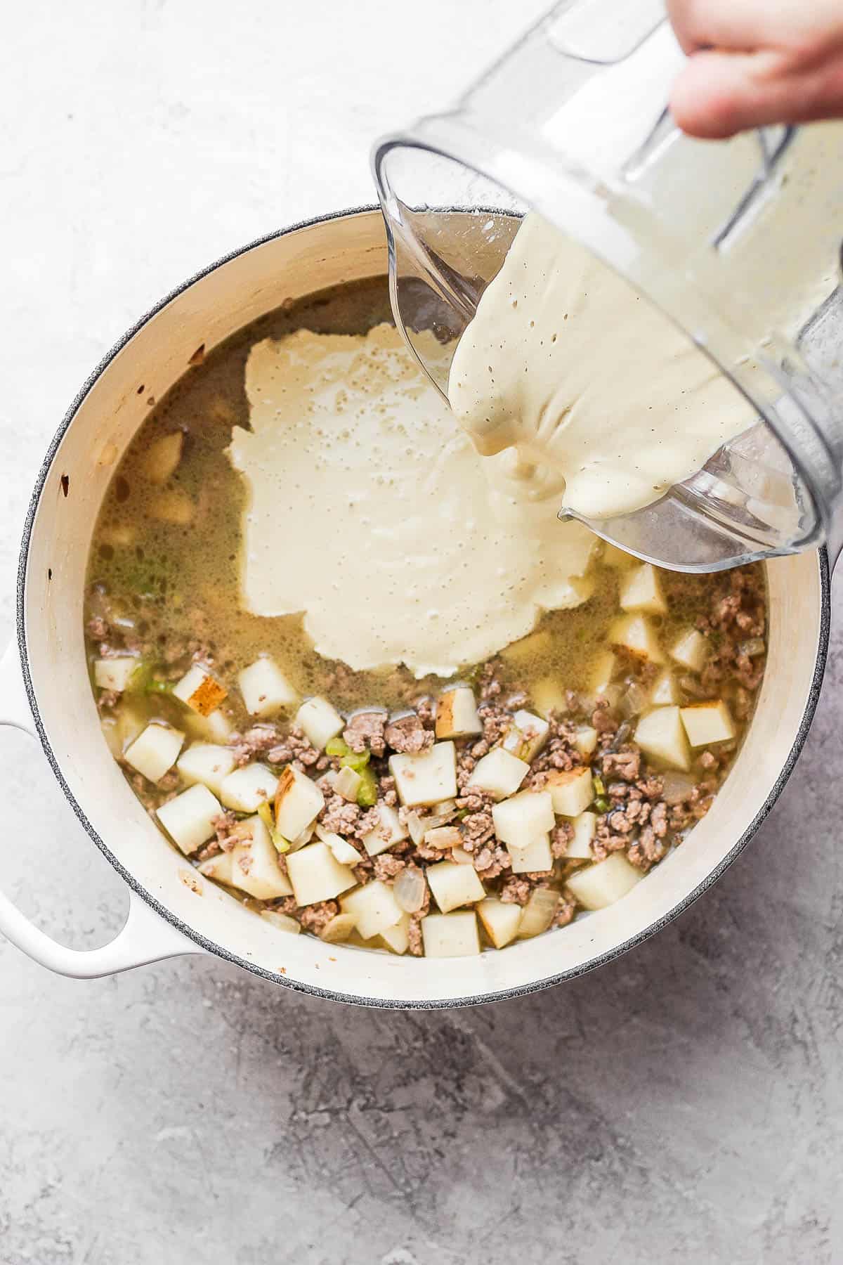 Cashew cream being poured into the dutch oven with the potatoes, broth and ground beef.