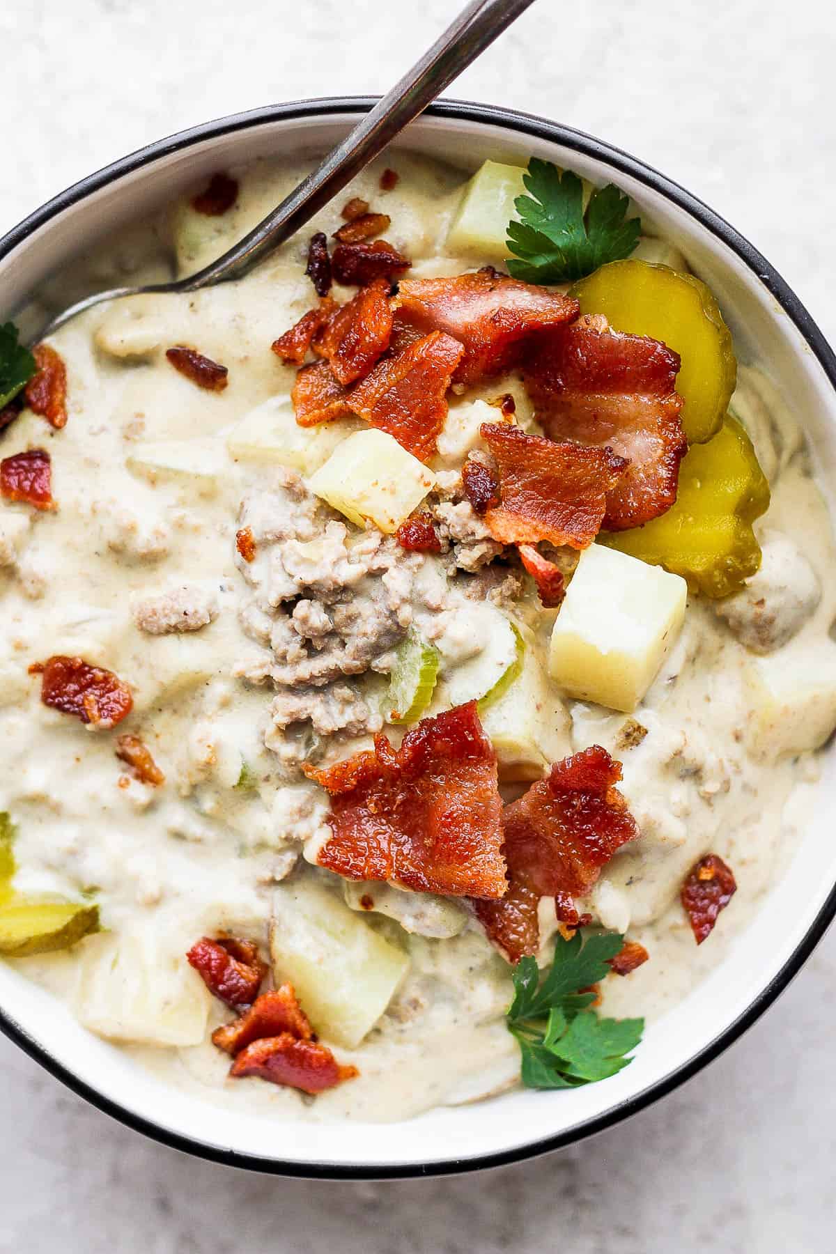 Bowl of cheeseburger soup garnished with parsley, pickles and bacon.