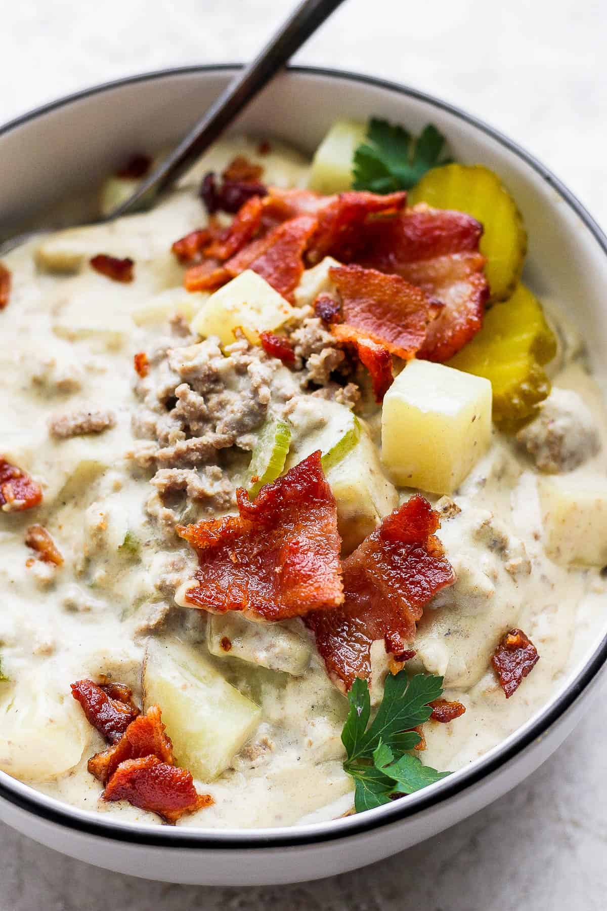 Bowl of cheeseburger soup garnished with parsley, pickles and bacon.