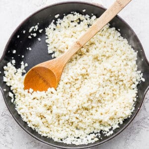 Coconut cauliflower rice in a cast iron skillet with a wooden spoon sticking out.