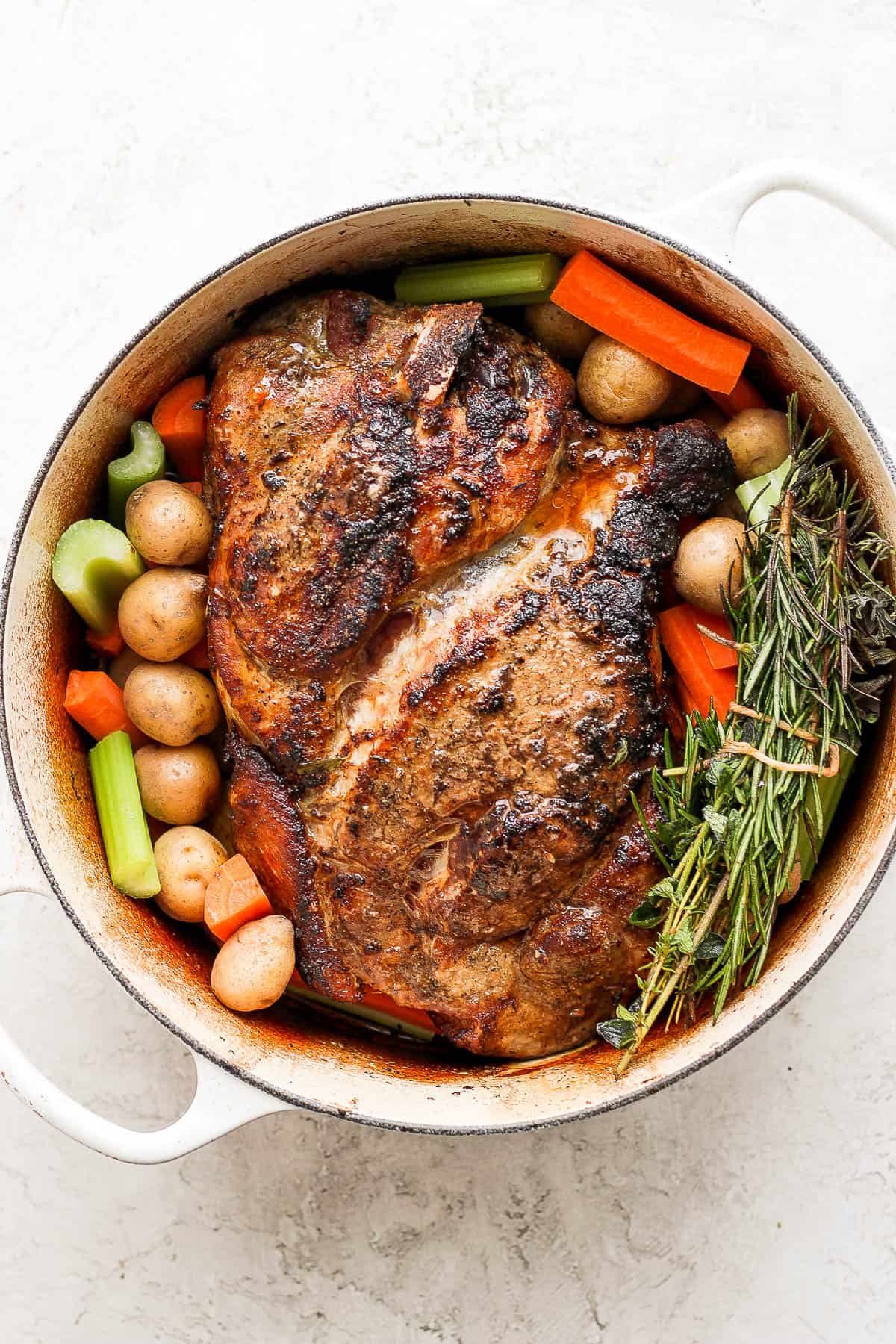 A pork roast recipe in a dutch oven with veggies all around it and an herb bundle. 