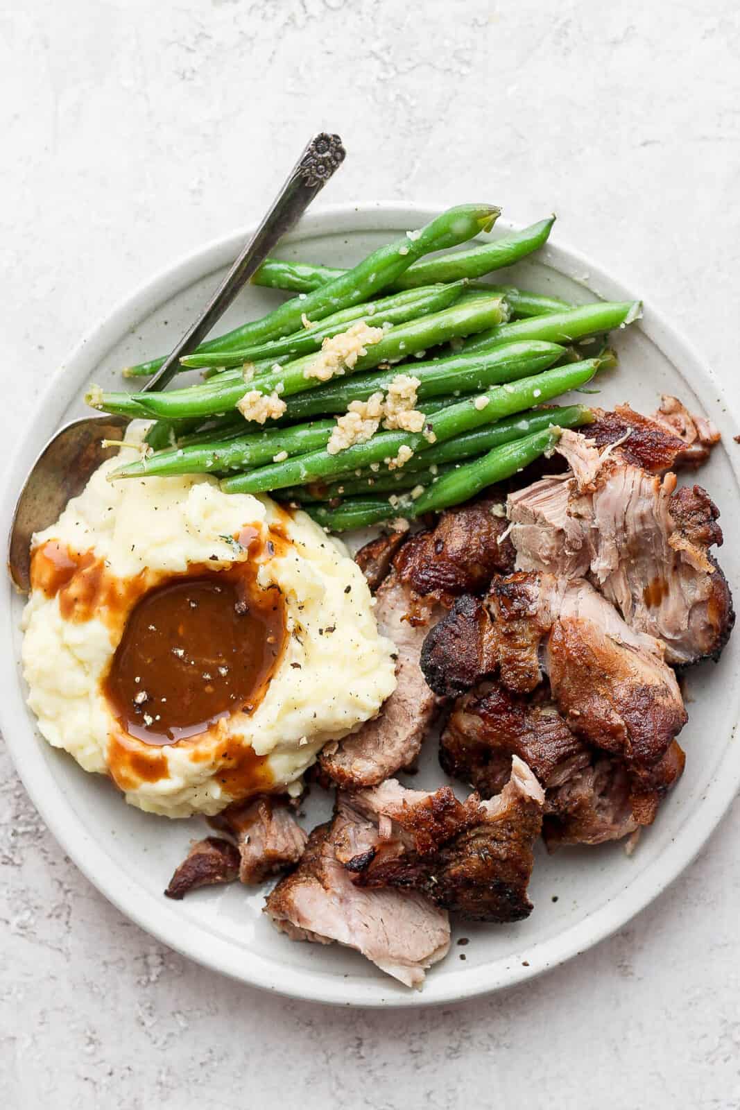 A plate of mashed potatoes with gravy, green beans, and pork roast.