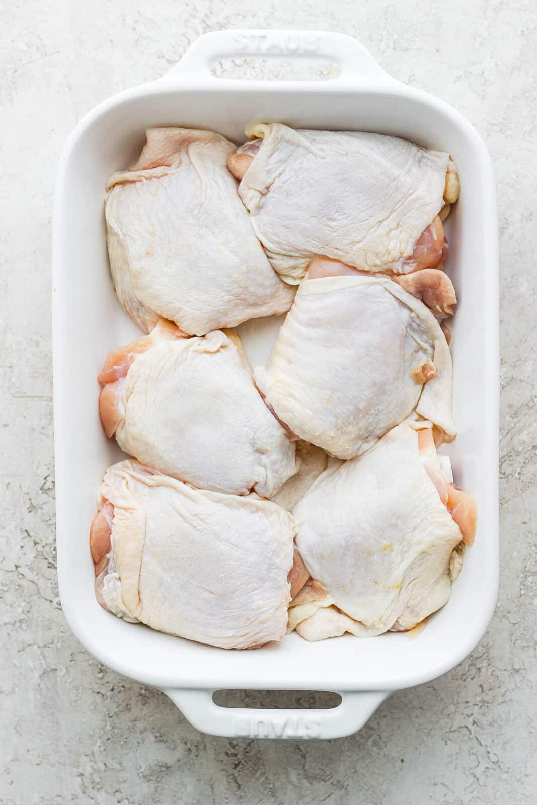 A white baking dish with 6 raw bone-in, skin-on chicken thighs. 