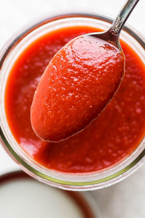 A spoon of homemade bbq sauce over a mason jar that is also full of bbq sauce.