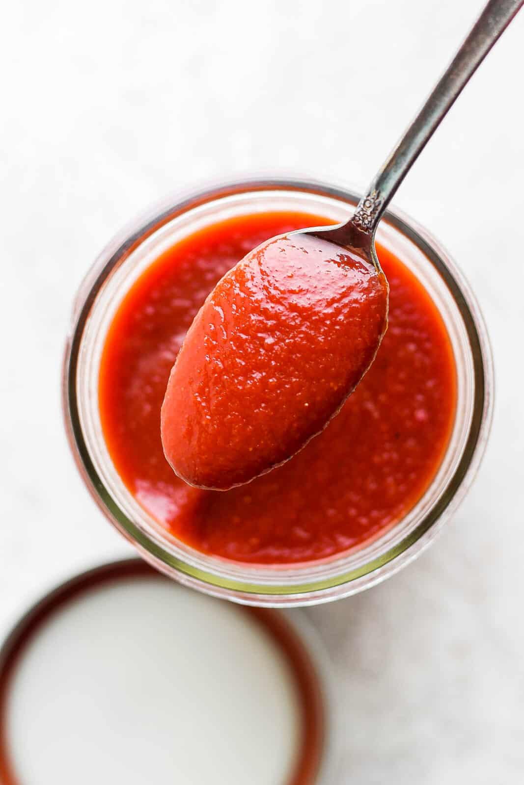 A spoon full of homemade bbq sauce hovering over a mason jar which is also full of bbq sauce.