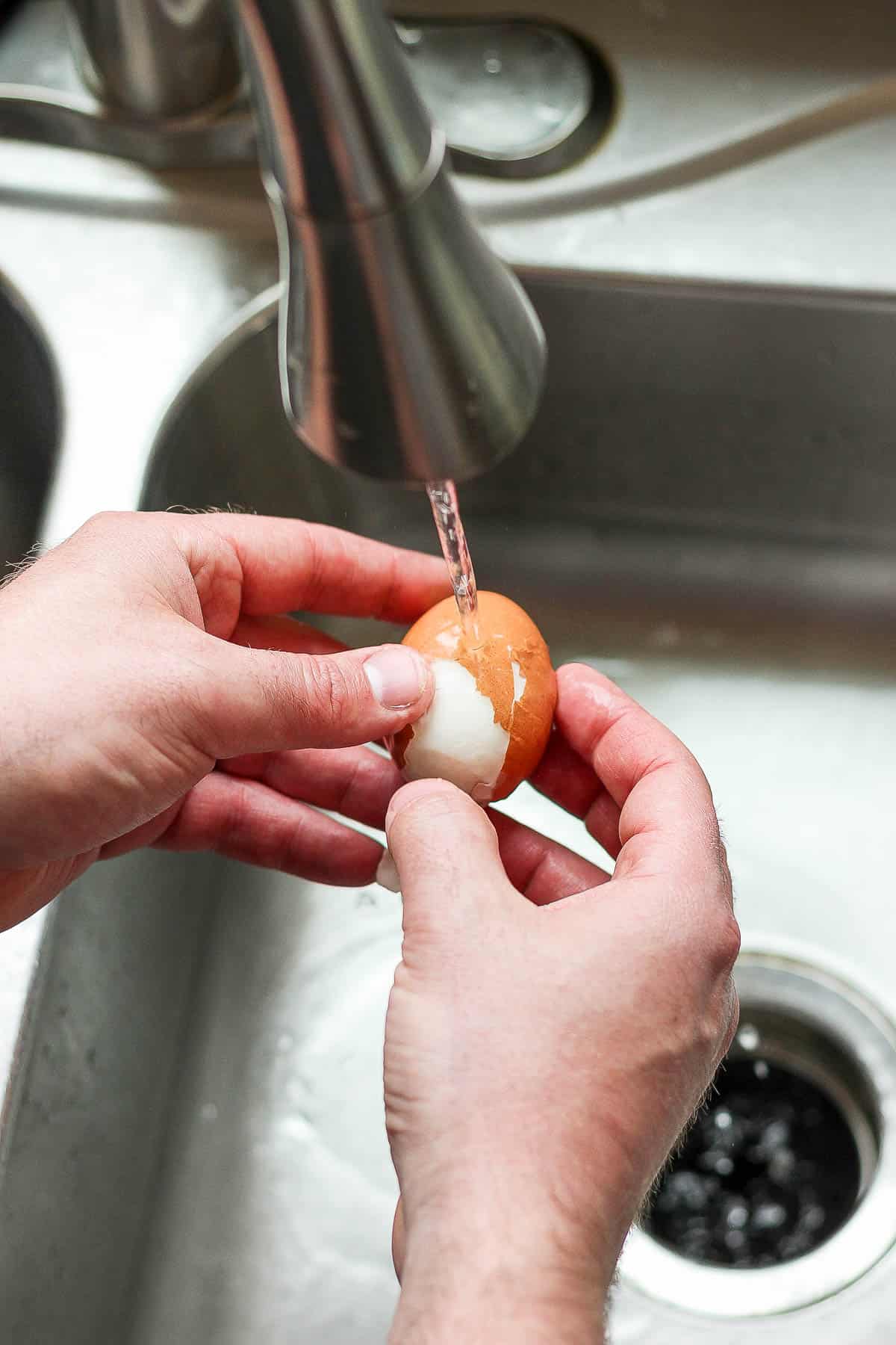 A hard boiled egg being peeled under running water.