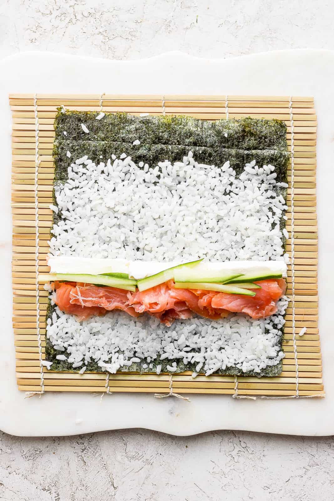 A nori sheet on top of a bamboo sheet with sushi rice spread on it and filling ingredients placed near the bottom.
