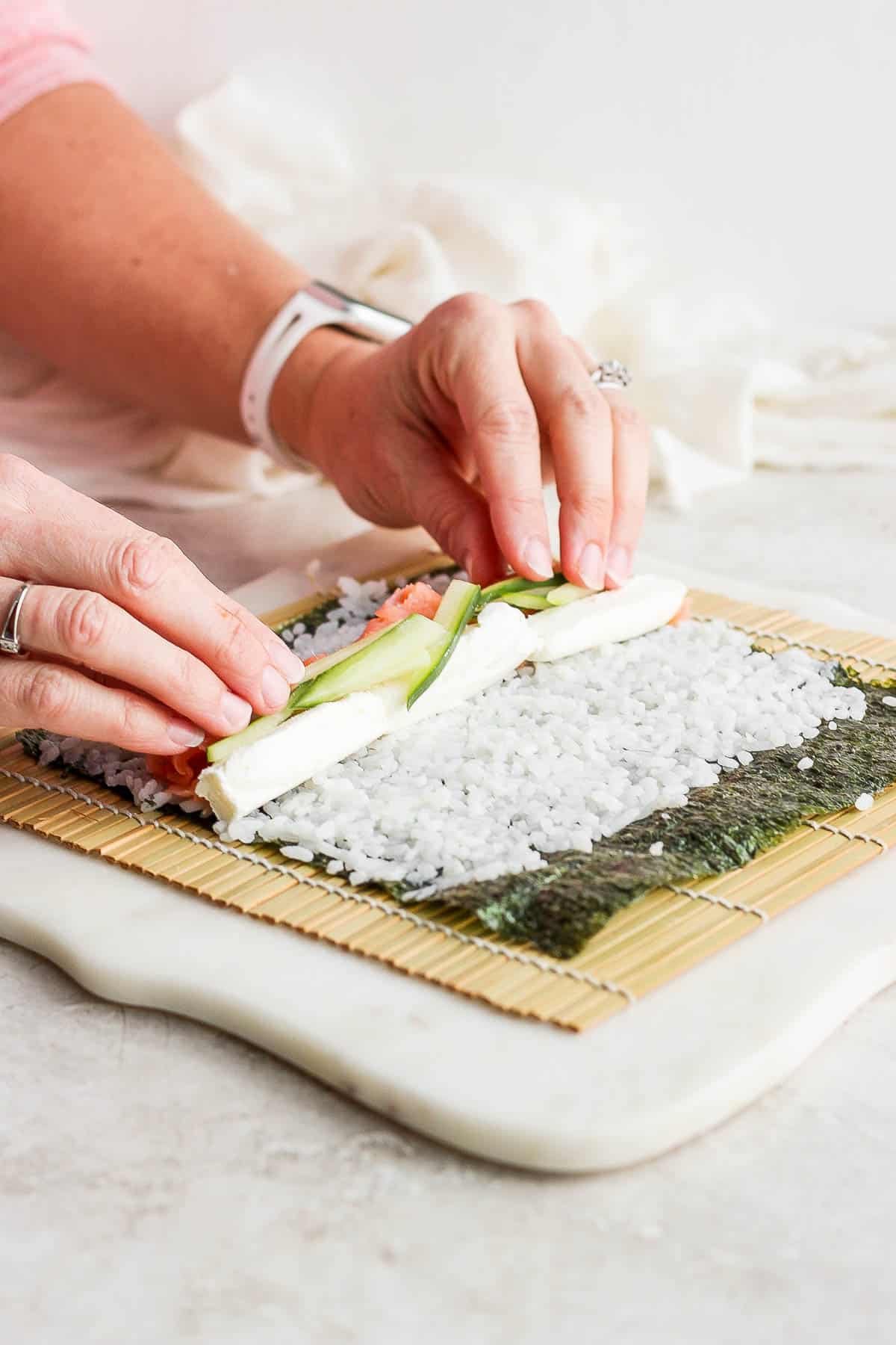 Two hands placing the filling ingredients towards the bottom of the nori sheet.