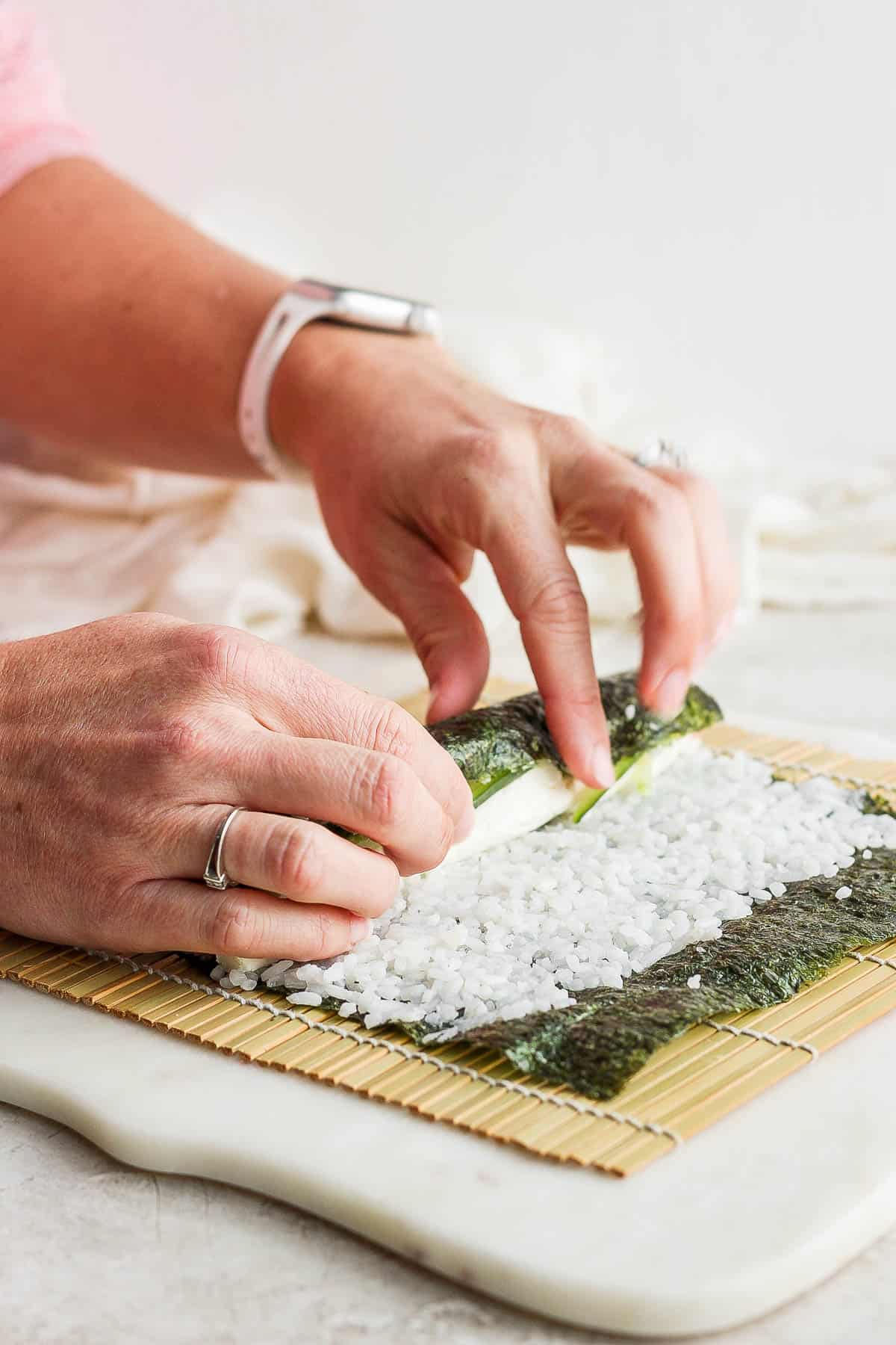 Two hands beginning to roll the nori sheet over the filling ingredients.