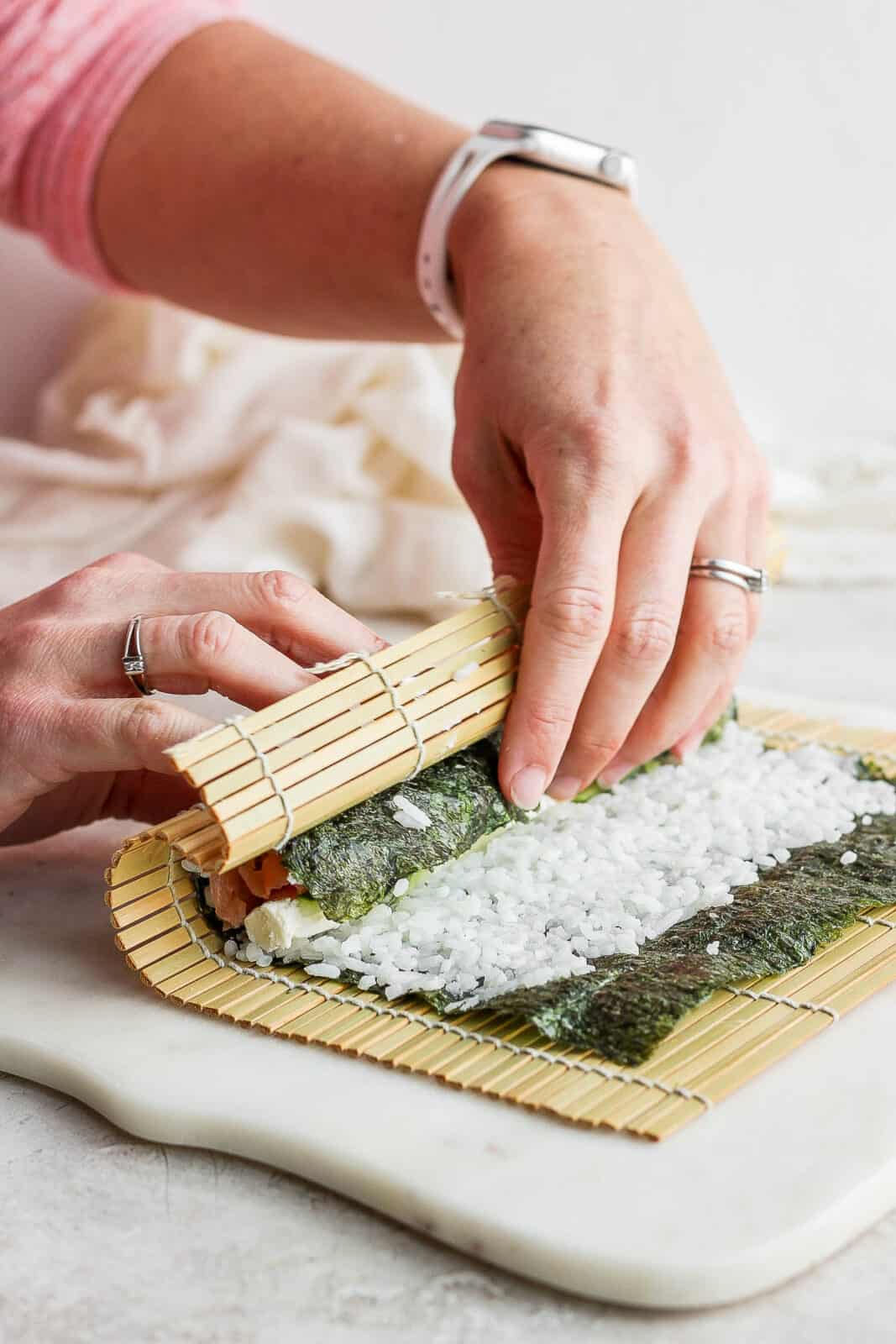 Two hands using the bamboo sheet to begin rolling the sushi roll.