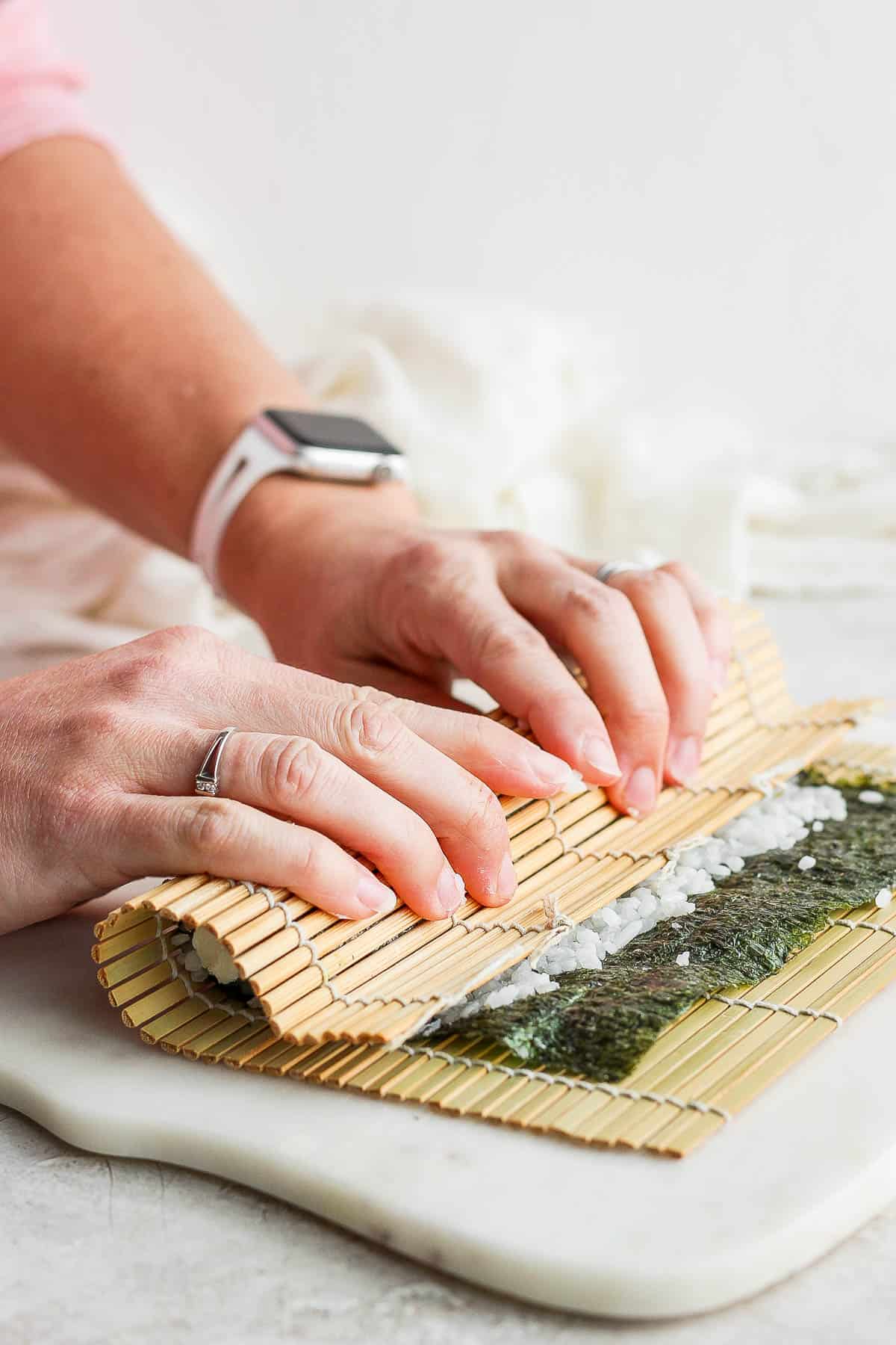Two hands tucking the nori sheet and bamboo sheet over the filling.