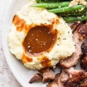 Close-up image of pork gravy on mashed potatoes with green beans and pork roast on the sides.