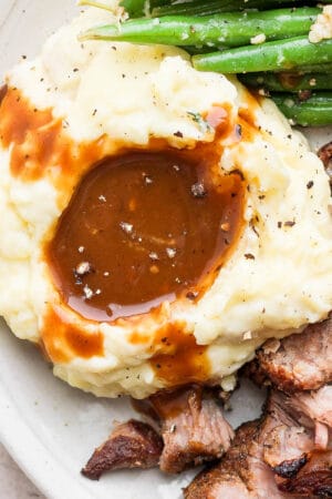 Close-up image of pork gravy on mashed potatoes with green beans and pork roast on the sides.