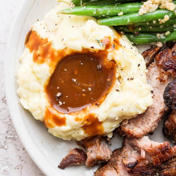 Close-up image of pork gravy on mashed potatoes with green beans and pork roast on the sides.