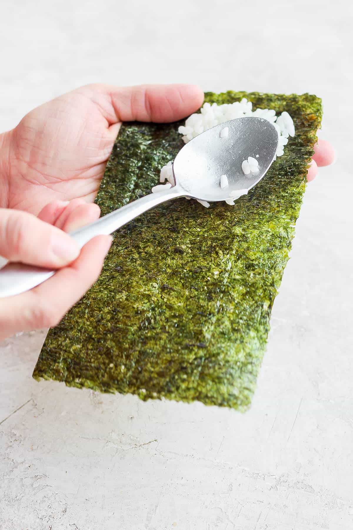 Someone using a spoon to spread sushi rice on half a nori sheet for a temaki sushi hand roll.