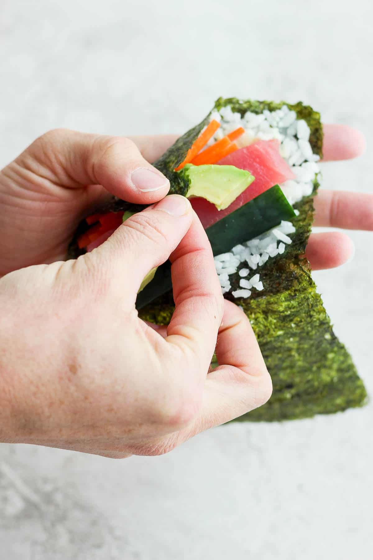 The nori sheet corner near the thumb being tucked across the filling ingredients to start forming a hand roll.