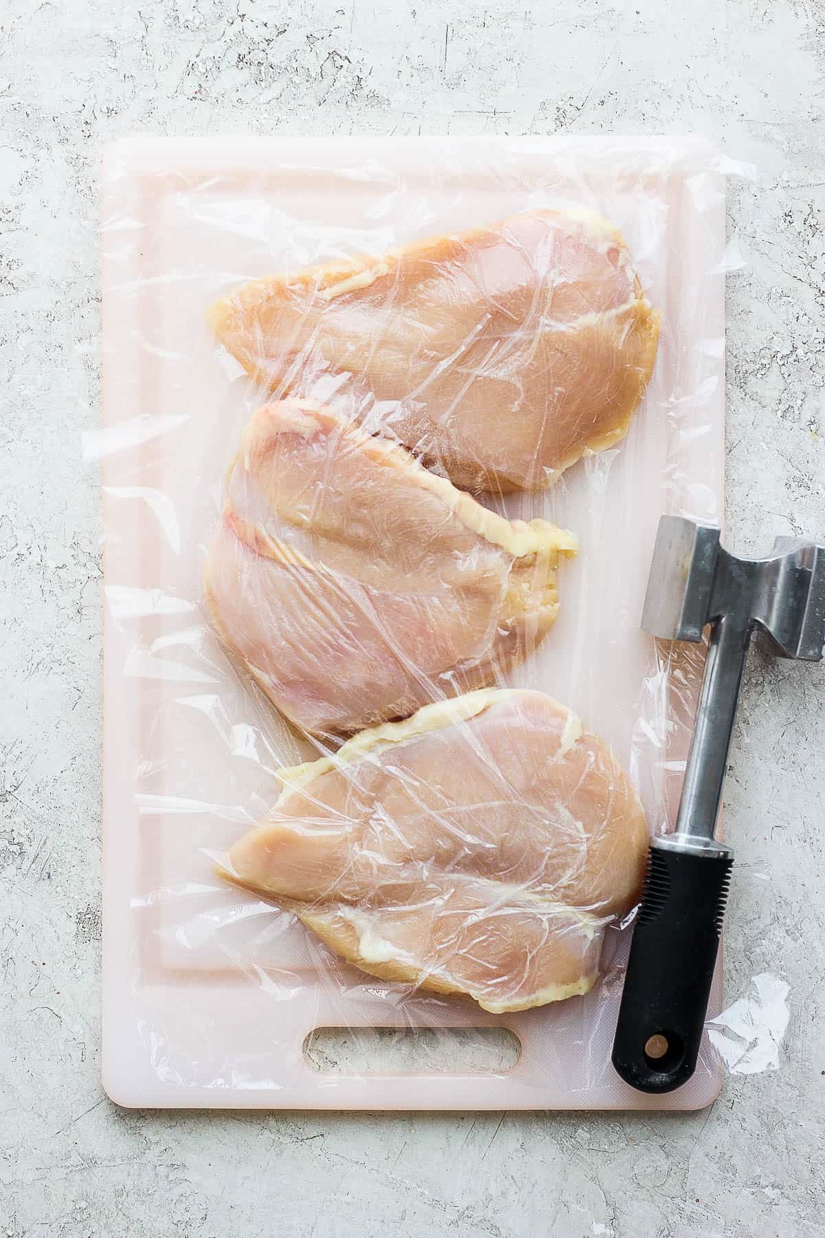 Raw chicken breasts on a cutting board with plastic wrap over them and a meat tenderizer.