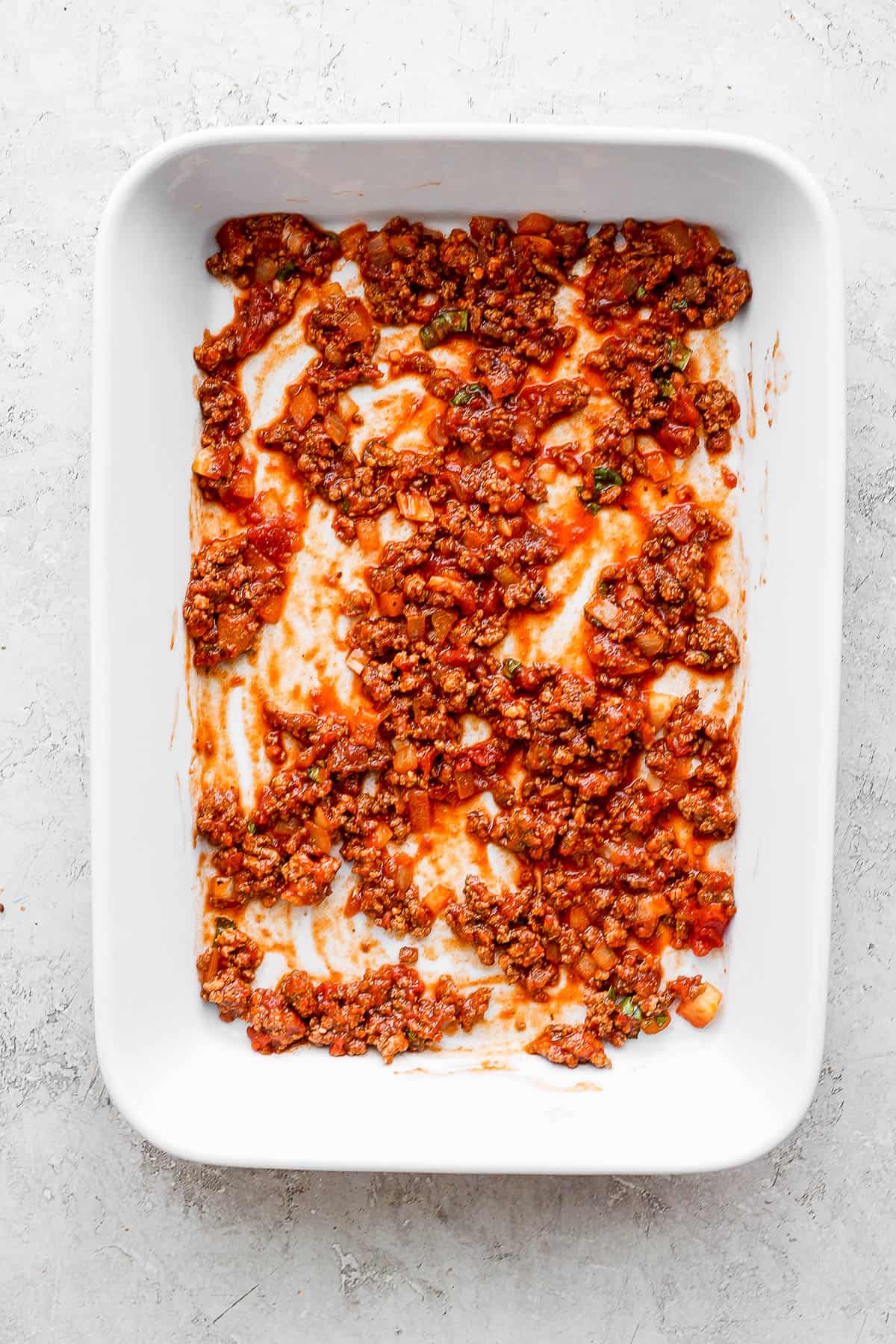 A small amount of marinara sauce with meat on the bottom of a white baking dish.