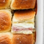 Several ham and cheese sliders in a white baking dish with one slider turned to see the inside.