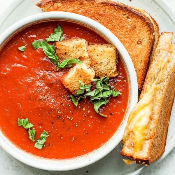 Bowl of creamy tomato basil soup with croutons on a plate with a grilled cheese sandwich.