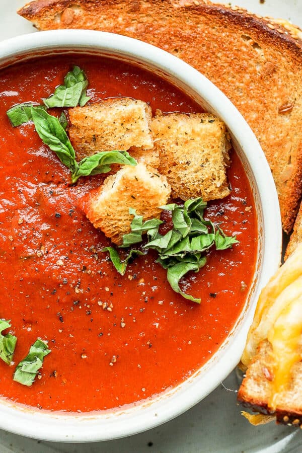 Bowl of creamy tomato basil soup with croutons on a plate with a grilled cheese sandwich.