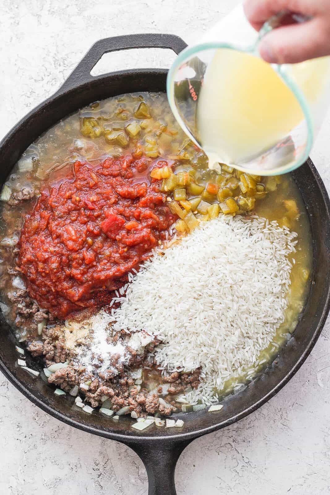 A cast iron skillet filled with ground beef, chile peppers, salsa and rice with someone pouring broth in.