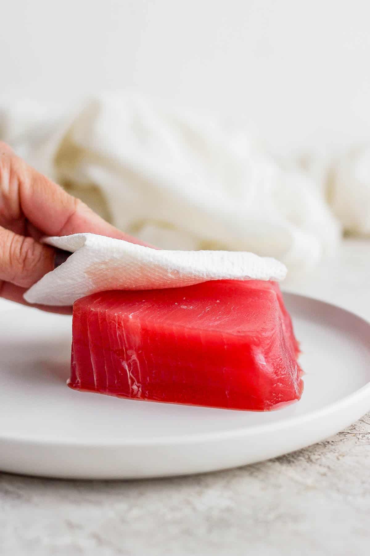A raw tuna steak on a white plate being pat dry with paper towel.