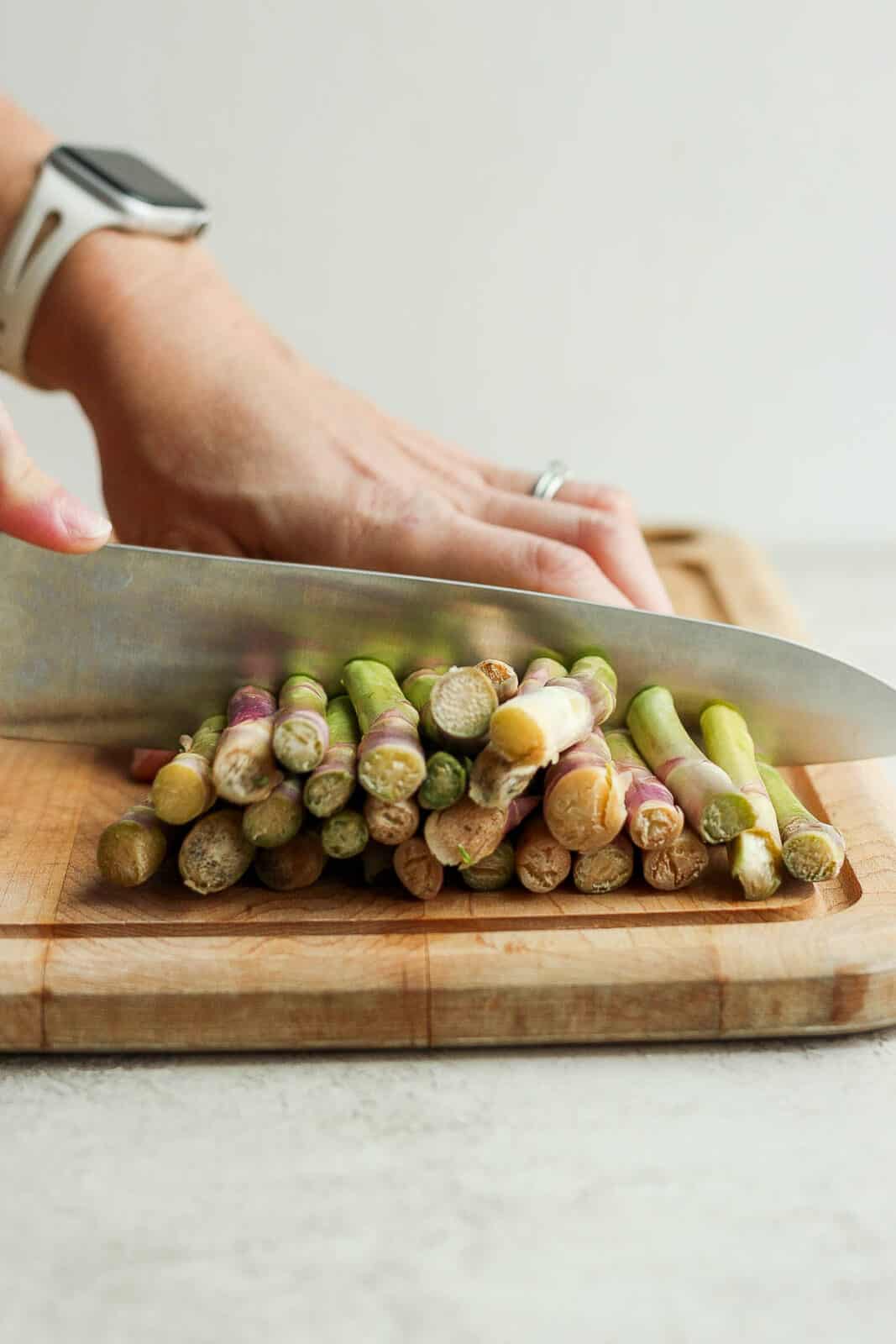 The ends of a bunch of asparagus being cut off with a knife on a wood cutting board.